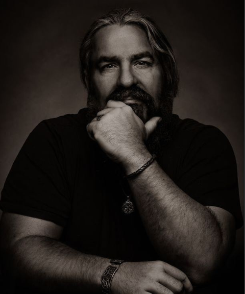 Neil Wallace artist bio profile photo Neil sits for a portrait in a photo studio. He looks directly down the lens while resting his chin on his left fist.